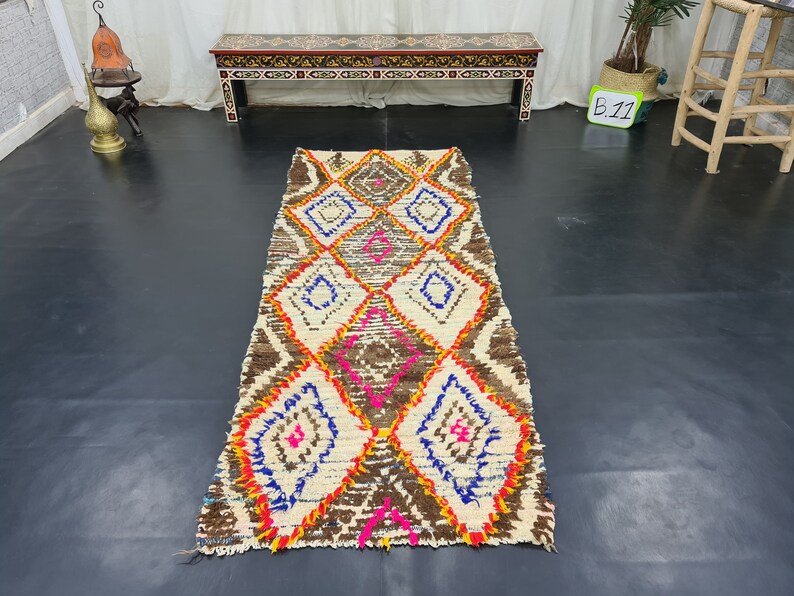 May include: A white, brown, orange, pink, and blue Moroccan rug with a diamond pattern. The rug is approximately 8 feet long and 2 feet wide.