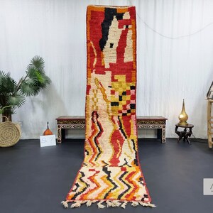 May include: A long, narrow, handwoven rug with a geometric pattern in shades of red, orange, yellow, white, and black. The rug has a fringe on both ends.