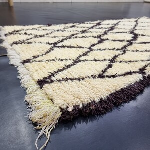 May include: A white and brown Moroccan rug with a geometric pattern. The rug has a fringe border and is made of wool.