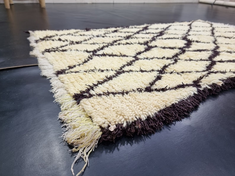 May include: A white and brown Moroccan rug with a geometric pattern. The rug has a fringe border and is made of wool.