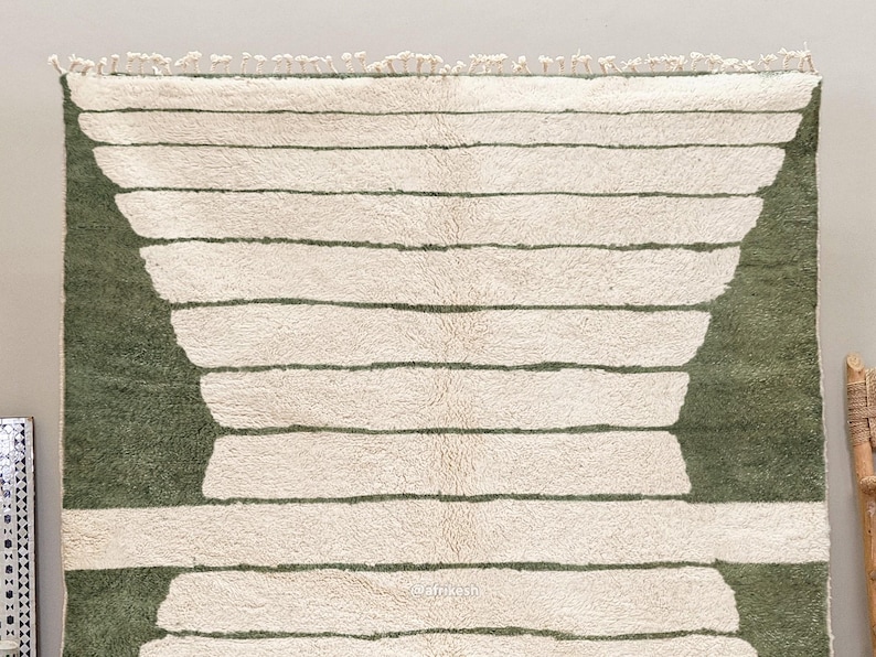 May include: A rectangular rug with a cream-colored background and horizontal olive green stripes. The rug has a geometric design with a fringe at the top. The rug's design features a series of horizontal stripes and a curved shape on each side.