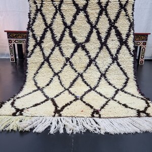 May include: A white and brown Moroccan rug with a diamond pattern and fringe. The rug is folded in half and is lying on a black floor.