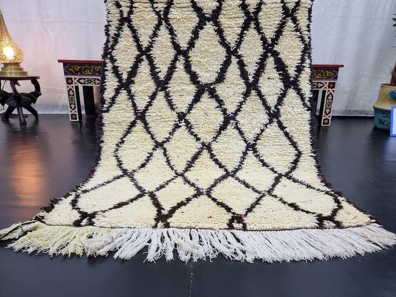 May include: A white and brown Moroccan rug with a diamond pattern and fringe. The rug is folded in half and is lying on a black floor.