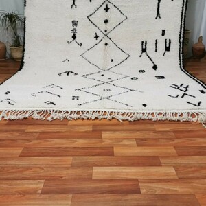 May include: A white and black Moroccan rug with a geometric pattern. The rug has a fringe border and is laid out on a wooden floor.