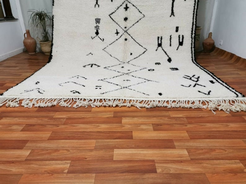 May include: A white and black Moroccan rug with a geometric pattern. The rug has a fringe border and is laid out on a wooden floor.