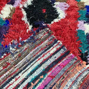 May include: A colorful, hand-woven rug with a mix of textures. The rug features a central section with a black and red design, surrounded by a border of red, white, and blue fringe.