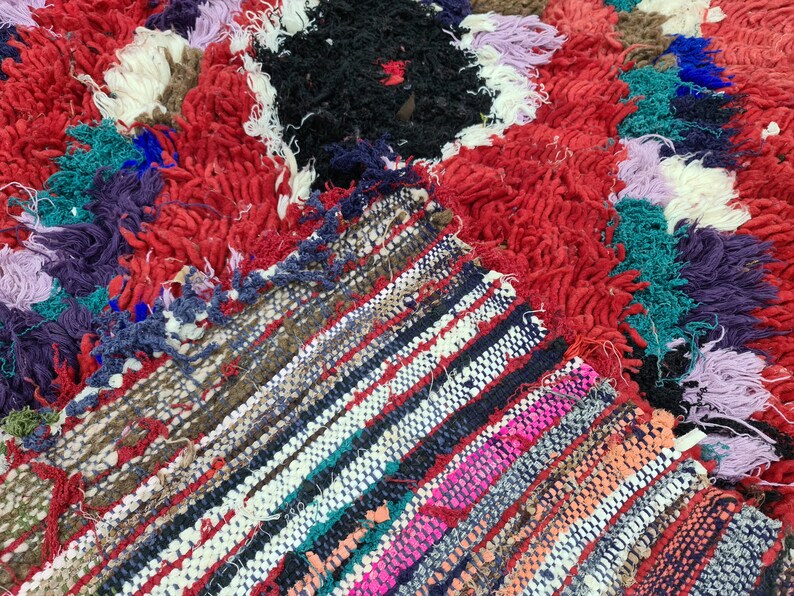 May include: A colorful, hand-woven rug with a mix of textures. The rug features a central section with a black and red design, surrounded by a border of red, white, and blue fringe.