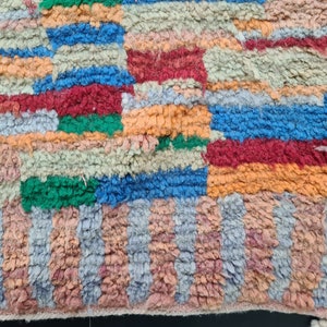 May include: A colorful, handwoven rug with a geometric pattern in shades of orange, red, blue, green, and gray. The rug has a textured, fuzzy surface.