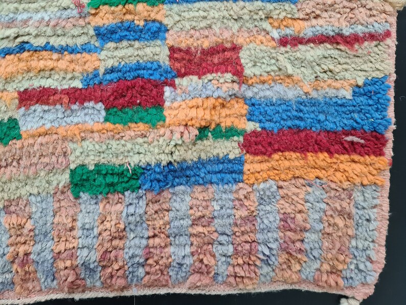 May include: A colorful, handwoven rug with a geometric pattern in shades of orange, red, blue, green, and gray. The rug has a textured, fuzzy surface.