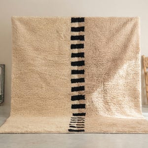 Puede incluir: Una alfombra beige grande con una franja vertical de formas rectangulares negras. La alfombra tiene una textura peluda y se muestra contra una pared neutra. Una silla tejida y una fuente decorativa están en el fondo.