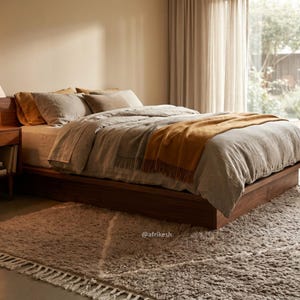 May include: A low-profile wooden platform bed frame with a light grey linen duvet, a mustard yellow throw blanket, and several pillows. A wooden nightstand with a lamp sits beside the bed, and a textured, cream-coloured rug with fringe lies below.