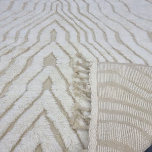 May include: A white and beige area rug with a geometric pattern and fringe. The rug is made of a soft, fluffy material.
