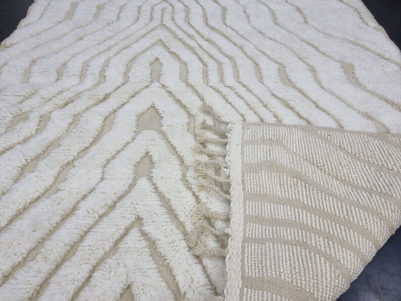 May include: A white and beige area rug with a geometric pattern and fringe. The rug is made of a soft, fluffy material.