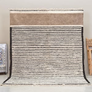 Puede incluir: Una alfombra rectangular grande con un patrón de rayas en negro y blanco roto. La sección superior presenta una banda sólida de color canela. La alfombra se exhibe sobre un suelo de color claro. Una pieza decorativa de mosaico y una silla de madera están en el fondo.