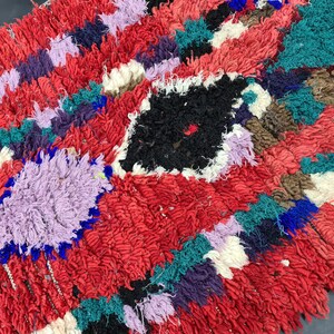 May include: A colorful, hand-woven rug with a red background and various geometric shapes in shades of purple, blue, green, and white.