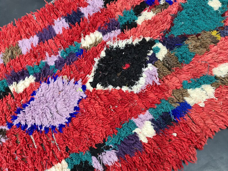 May include: A colorful, hand-woven rug with a red background and various geometric shapes in shades of purple, blue, green, and white.