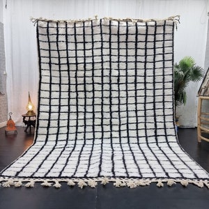 May include: A large, rectangular rug with a grid pattern of black lines on a white background. The rug has a thick, textured pile and a fringe of tassels along the bottom edge. The rug is displayed indoors.
