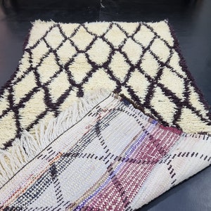 May include: A white and brown Moroccan rug with a diamond pattern. The rug is folded in half, revealing the back side, which is a multi-colored woven pattern.