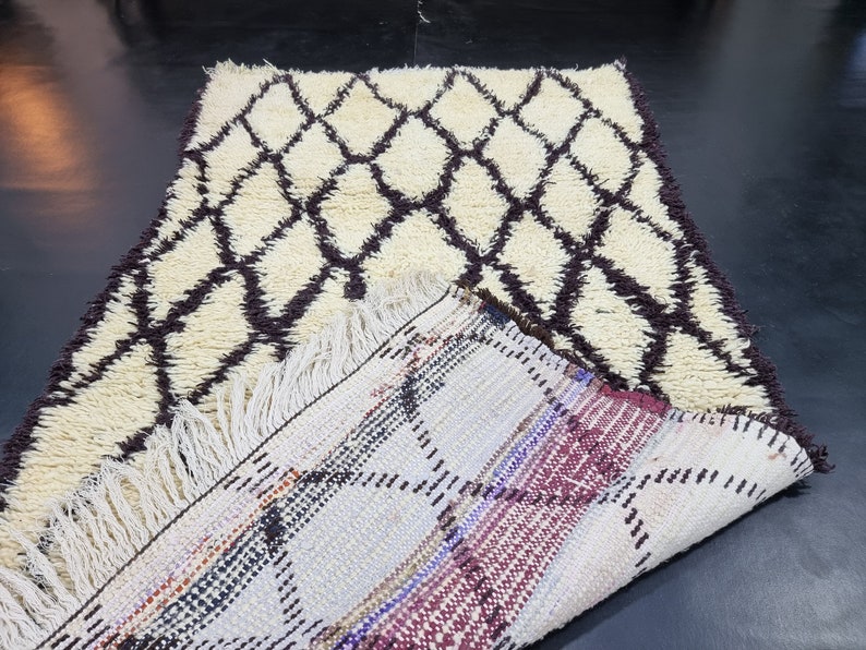 May include: A white and brown Moroccan rug with a diamond pattern. The rug is folded in half, revealing the back side, which is a multi-colored woven pattern.