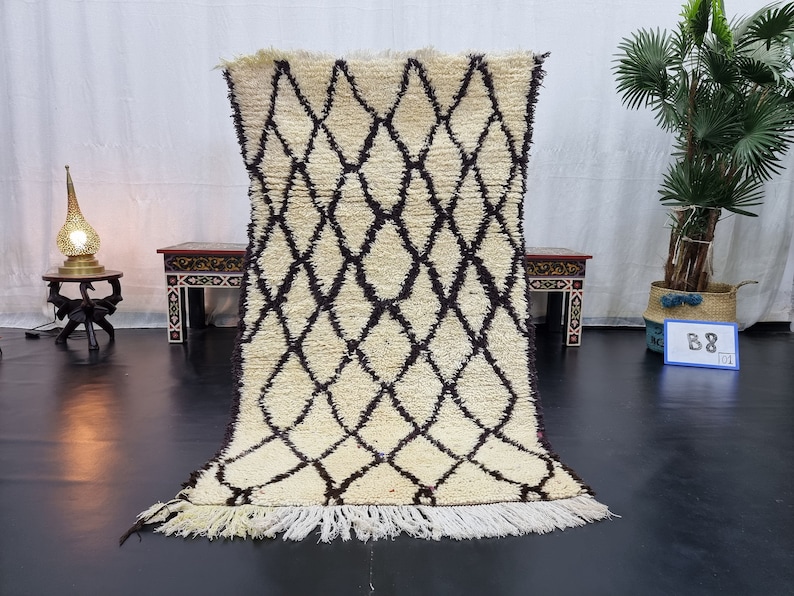 May include: A white and black Moroccan rug with a diamond pattern. The rug has a fringe border and is displayed on a black floor.