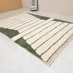 May include: A rectangular rug with a geometric design. The rug features a green border and a pattern of off-white rectangular shapes. The rug has a fringe on one end and is made of a soft, textured material.
