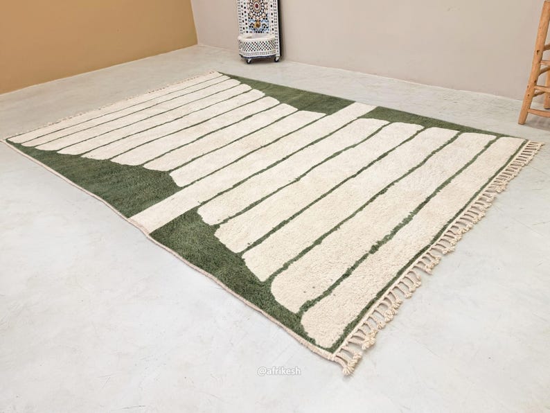 May include: A rectangular rug with a geometric design. The rug features a green border and a pattern of off-white rectangular shapes. The rug has a fringe on one end and is made of a soft, textured material.