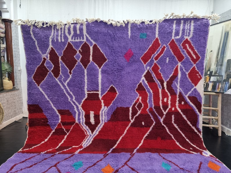 May include: A purple and red Moroccan rug with a geometric pattern. The rug has a fringe border and is made of wool.