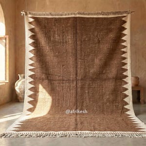 May include: A large, rectangular brown and cream-colored rug with a geometric zigzag border. The rug has a fringed edge and is displayed in a room with natural light. The rug is made of a woven material.