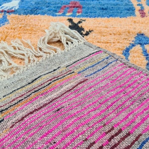 May include: Close-up of a woven rug with a fringe. The rug has a gray background with pink, blue, and orange stripes.