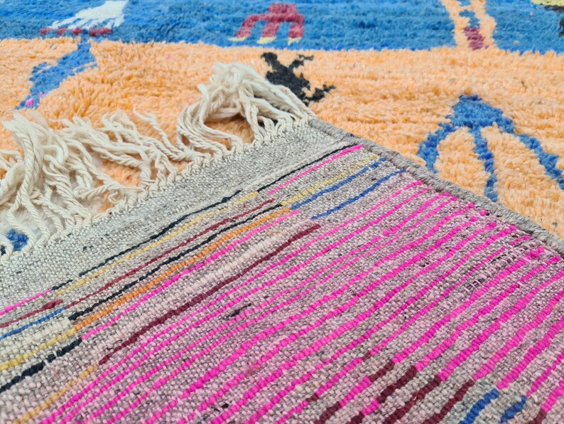 May include: Close-up of a woven rug with a fringe. The rug has a gray background with pink, blue, and orange stripes.