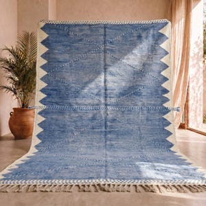 Puede incluir: Una alfombra grande, rectangular, azul y blanca con un diseño geométrico. La alfombra presenta un centro azul claro con bordes en zigzag blancos y patrones de diamantes. Tiene un borde con flecos y se exhibe en interiores.