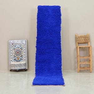 Op de afbeelding: Een lang, blauw, pluche vloerkleed met een witte rand. Het vloerkleed ligt plat op een lichtgrijze vloer.