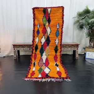 May include: A long, rectangular, orange Moroccan rug with a diamond pattern in shades of red, blue, green, and white. The rug has a fringe border and is laid out on a black floor.