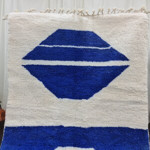 Puede incluir: Una alfombra marroqu&iacute; blanca y azul con un dise&ntilde;o geom&eacute;trico. La alfombra tiene un fondo blanco con una forma de diamante azul en el centro. La forma de diamante tiene tres rayas blancas a trav&eacute;s de ella. La parte inferior de la alfombra es de color azul s&oacute;lido.