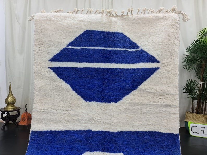 Puede incluir: Una alfombra marroqu&iacute; blanca y azul con un dise&ntilde;o geom&eacute;trico. La alfombra tiene un fondo blanco con una forma de diamante azul en el centro. La forma de diamante tiene tres rayas blancas a trav&eacute;s de ella. La parte inferior de la alfombra es de color azul s&oacute;lido.