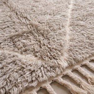 May include: A thick, high-pile area rug in a light beige and cream colour palette. The rug features a subtle, large-scale diamond pattern formed by smooth, flat-woven lines against the shaggy texture. The edge is finished with braided fringe.