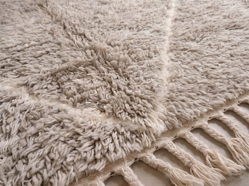 May include: A thick, high-pile area rug in a light beige and cream colour palette. The rug features a subtle, large-scale diamond pattern formed by smooth, flat-woven lines against the shaggy texture. The edge is finished with braided fringe.