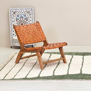May include: A wooden chair with a woven leather seat and back. The chair is brown and sits on a green and white patterned rug. A decorative mosaic cabinet is in the background.