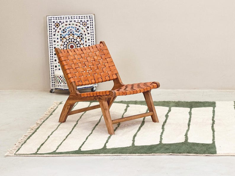 May include: A wooden chair with a woven leather seat and back. The chair is brown and sits on a green and white patterned rug. A decorative mosaic cabinet is in the background.