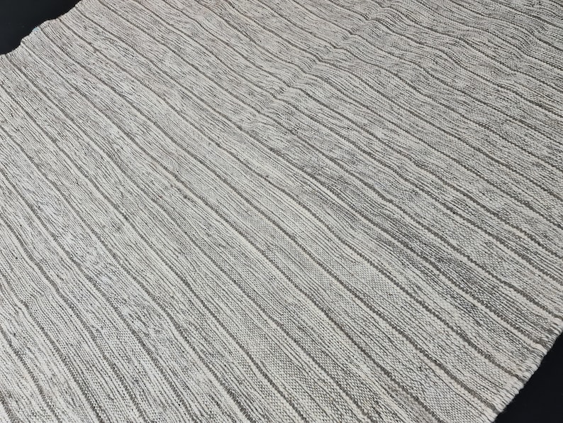 Puede incluir: Una alfombra blanca tejida con un patr&oacute;n texturizado a rayas. La alfombra tiene un brillo sutil y una textura ligeramente rugosa.