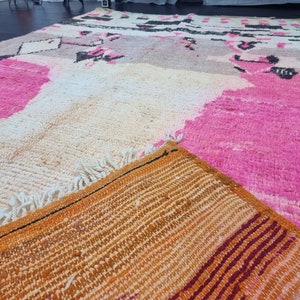 May include: A close-up of a colorful Moroccan rug with pink, white, and orange hues. The rug has a woven texture and a fringe border.