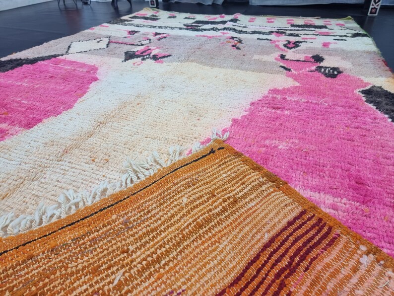 May include: A close-up of a colorful Moroccan rug with pink, white, and orange hues. The rug has a woven texture and a fringe border.