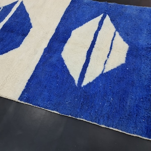 Puede incluir: Una alfombra geom&eacute;trica con estampado blanco y azul con flecos. La alfombra tiene un fondo blanco con formas de diamantes azules.