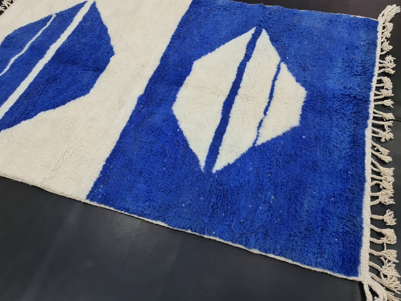 Puede incluir: Una alfombra geom&eacute;trica con estampado blanco y azul con flecos. La alfombra tiene un fondo blanco con formas de diamantes azules.