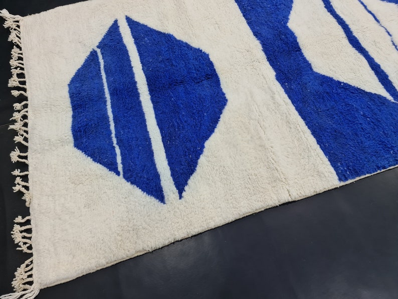 Puede incluir: Una alfombra blanca con un patr&oacute;n geom&eacute;trico azul. La alfombra tiene un borde de flecos.