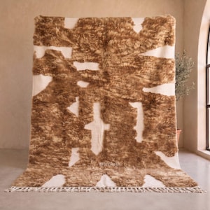 May include: A rectangular area rug featuring a shaggy, high-pile texture with an abstract pattern of irregular brown patches against a cream-colored background. The rug includes a fringe detail along the bottom edge and the text @afrikesh.