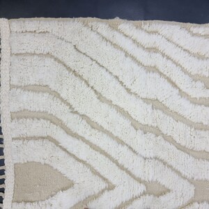 May include: A white and beige rug with a wavy, abstract pattern. The rug has a fringe border.
