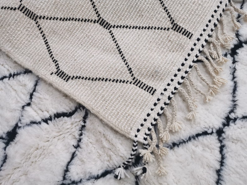 May include: Two white rugs with black geometric patterns. The top rug has a fringe border and a diamond pattern. The bottom rug has a more intricate pattern.