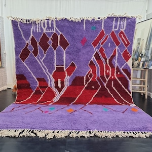 May include: A purple and red Moroccan rug with a geometric pattern. The rug has a fringe border and is made of wool.