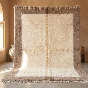 Puede incluir: Una alfombra rectangular de pelo largo con un campo central de color crema enmarcado por un borde ancho y texturizado en un tono marrón topo. La alfombra incluye una línea vertical de color crema en el centro y flecos decorativos.